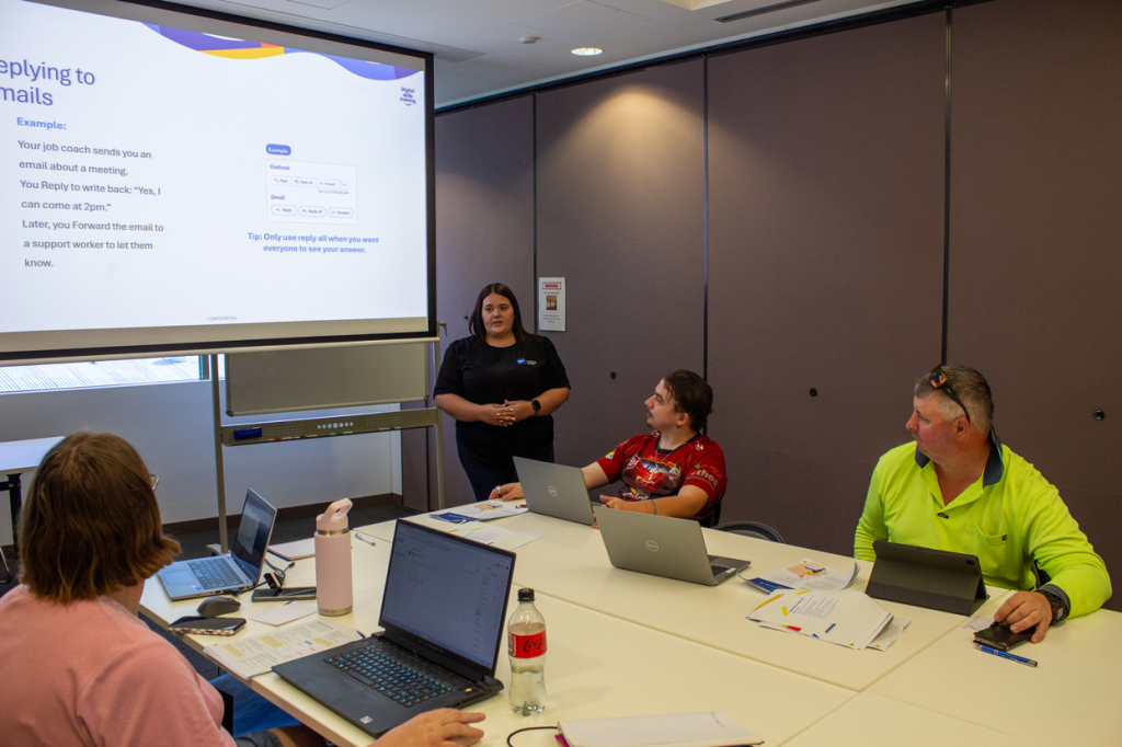 Emily, the Digital Skills Trainer, is teaching a workshop with three participants (one woman, two men) around a large table. Each person has a device in front of them and they are all looking towards the projector screen.